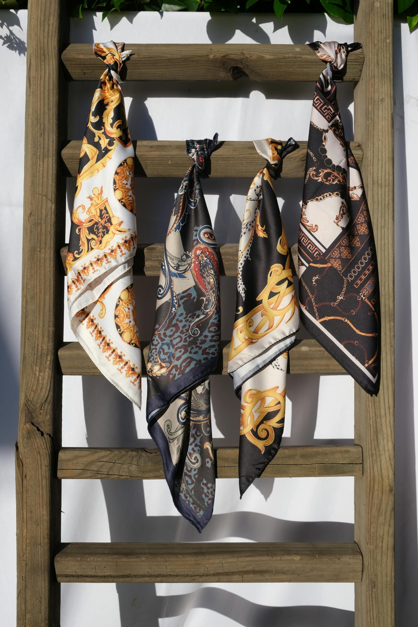 Four intricately designed silk scarves hang gracefully on a wooden ladder against a sunny backdrop.