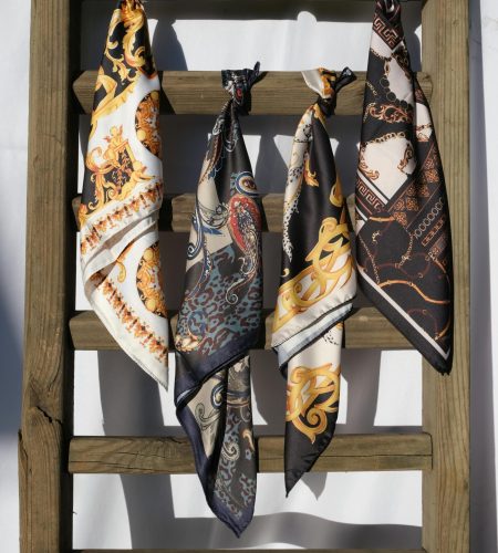Four intricately designed silk scarves hang gracefully on a wooden ladder against a sunny backdrop.